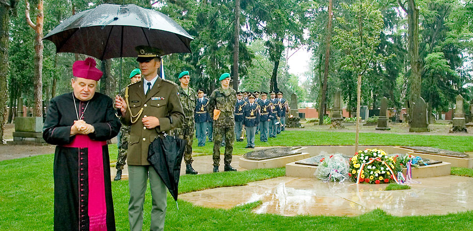 Církevní hodnostář stojící pod deštníkem, který drží voják.